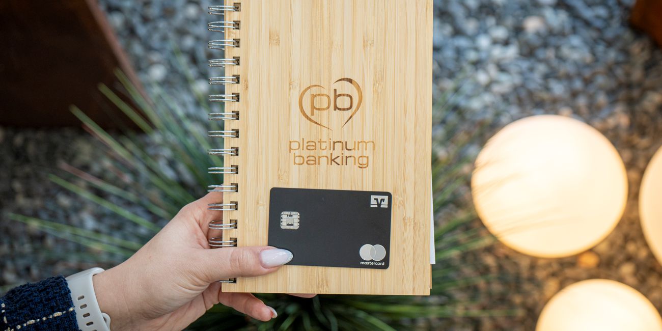 Eine Hand hält eine schwarze Kreditkarte auf einem hölzernen Hintergrund mit platinum Banking Logo.
