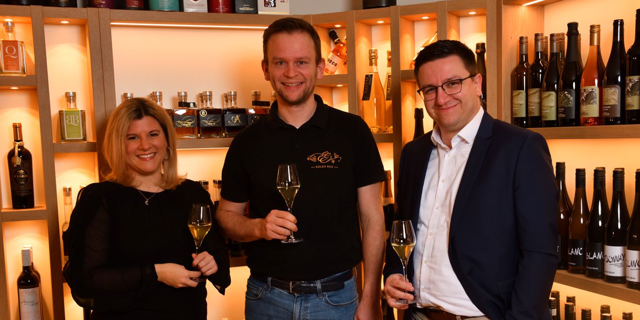 Sonja und Hannes Wenger mit Private Banking Berater Julian Bachert bei einem Glas Wein, im Hintergrund sind unterschiedliche Getränkeflaschen ausgestellt.