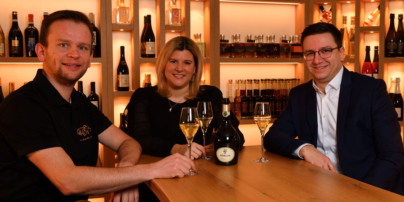 Hannes und Sonja Wenger mit Private Banking Berater Julian Bachert bei einem Glas Wein, im Hintergrund sind unterschiedliche Getränkeflaschen ausgestellt.