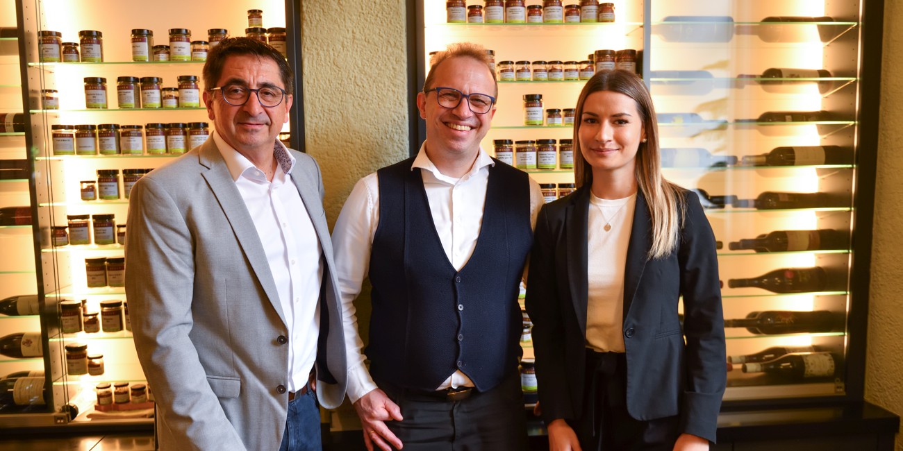 Drei Personen stehen nebeneinander in einem Feinkostgeschäft vor einem Regal mit Gläsern und Weinflaschen. Zwei Männer im Anzug bzw. mit Weste und Hemd, eine Frau im Blazer.