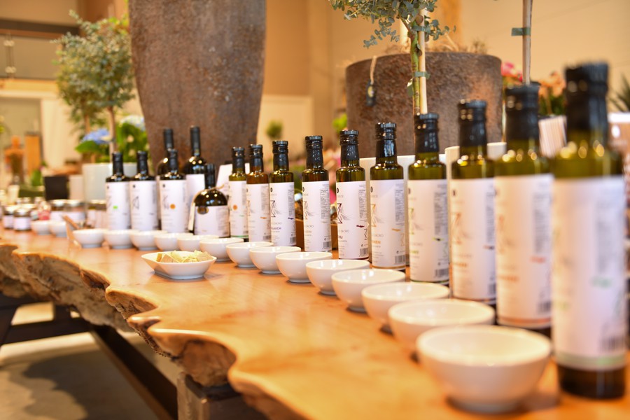 Langer Holztisch mit einer Reihe an Olivenölflaschen und kleinen weißen Probierschalen, dekorativ arrangiert.