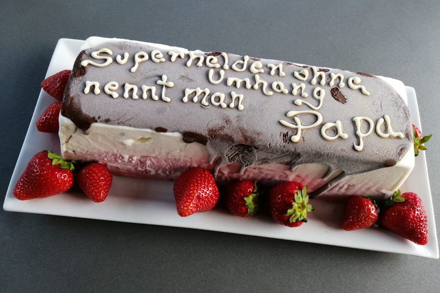 Eine kastenförmige Eistorte umrahmt mit Erdbeeren beschriftet mit: Superhelden ohne Umhang nennt man Papa.