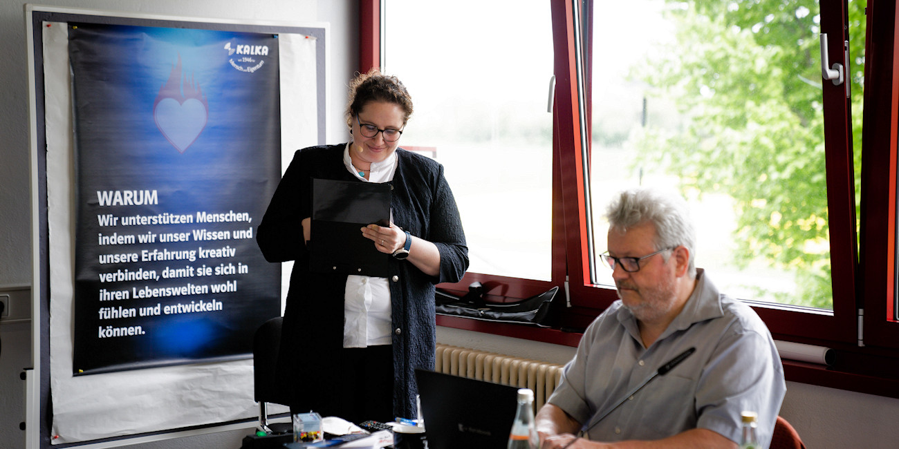 Eva Heißwolf schreibt auf einem Klemmbrett und Reinhard Schweizer am Laptop, im Hintergrund ein Plakat mit der Aufschrift: WARUM – Wir unterstützen Menschen, indem wir unser Wissen und unsere Erfahrung kreativ verbinden, damit sie sich in ihren Lebenswelten wohl fühlen und entwickeln können.