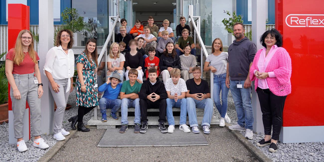 Schülerinnen und Schüler einer achten Klasse aus der Schulpartnerschaft mit den Verantwortlichen aus dem Unternehmen sitzen vor dem Eingang der Firma Reflexa.