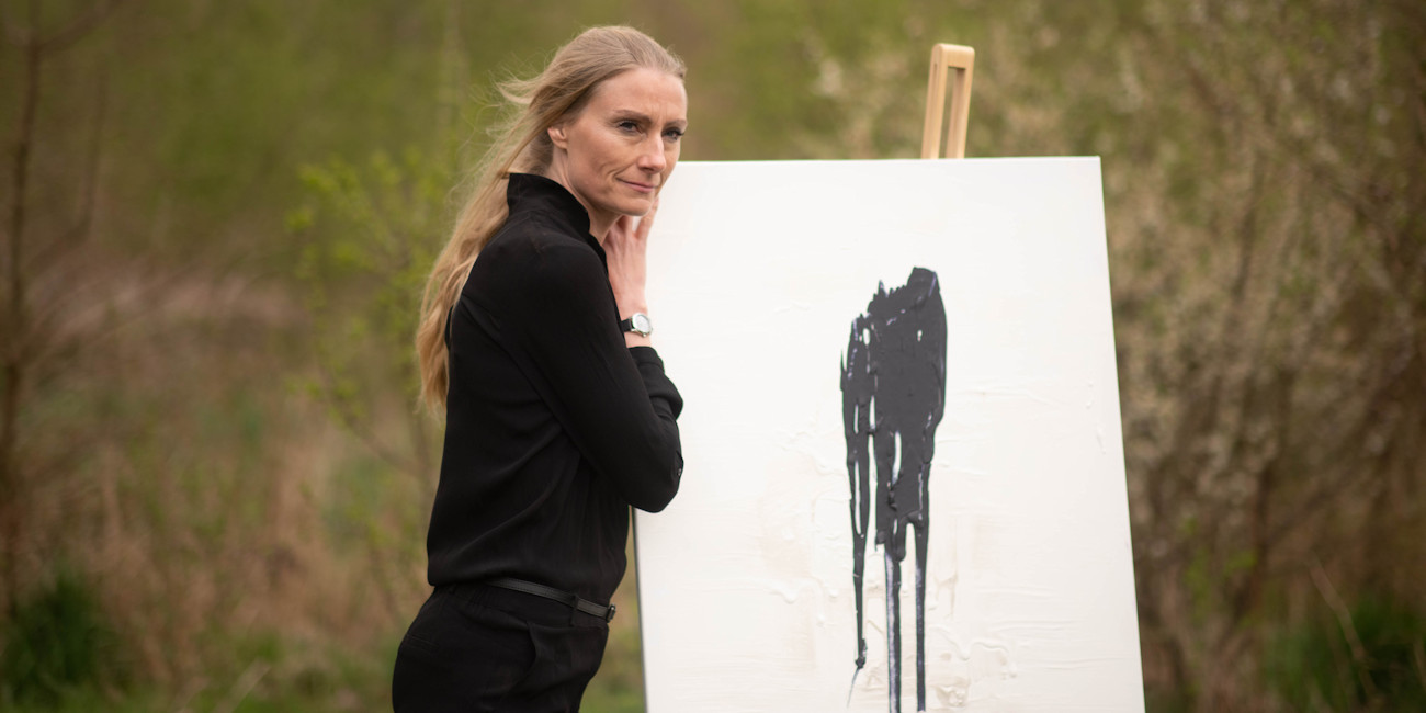 Stefanie Rau steht neben einer Leinwand mit einem Gemälde in der Natur. 
