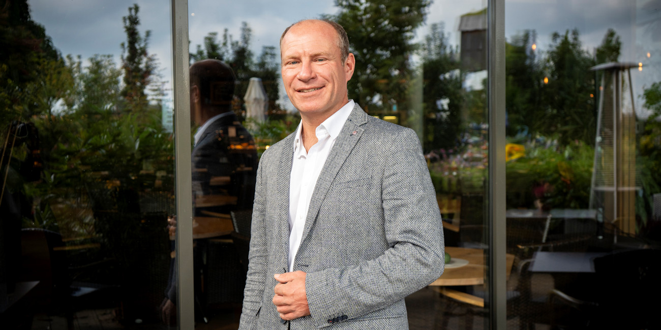 Jürgen Wohlhüter steht vor einer Fensterfront, in der sich ein Garten spiegelt.