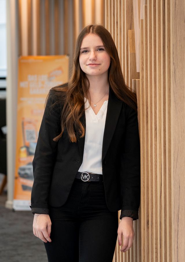 Maria Schmid, Assistenz PlatinumTeam lehnt an einer Holzwand mit VR-Bank Logo.