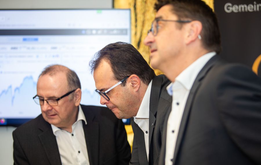 3 Berater Private Banking in Anzügen schauen gemeinsam in eine Richtung. Im Hintergrund ist ein großer Bildschirm mit einem Chart.